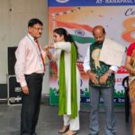 Ceremony on a stage where a woman pins a medal on a man as others watch another man stands nearby and a woman in a saree holds a bouquet